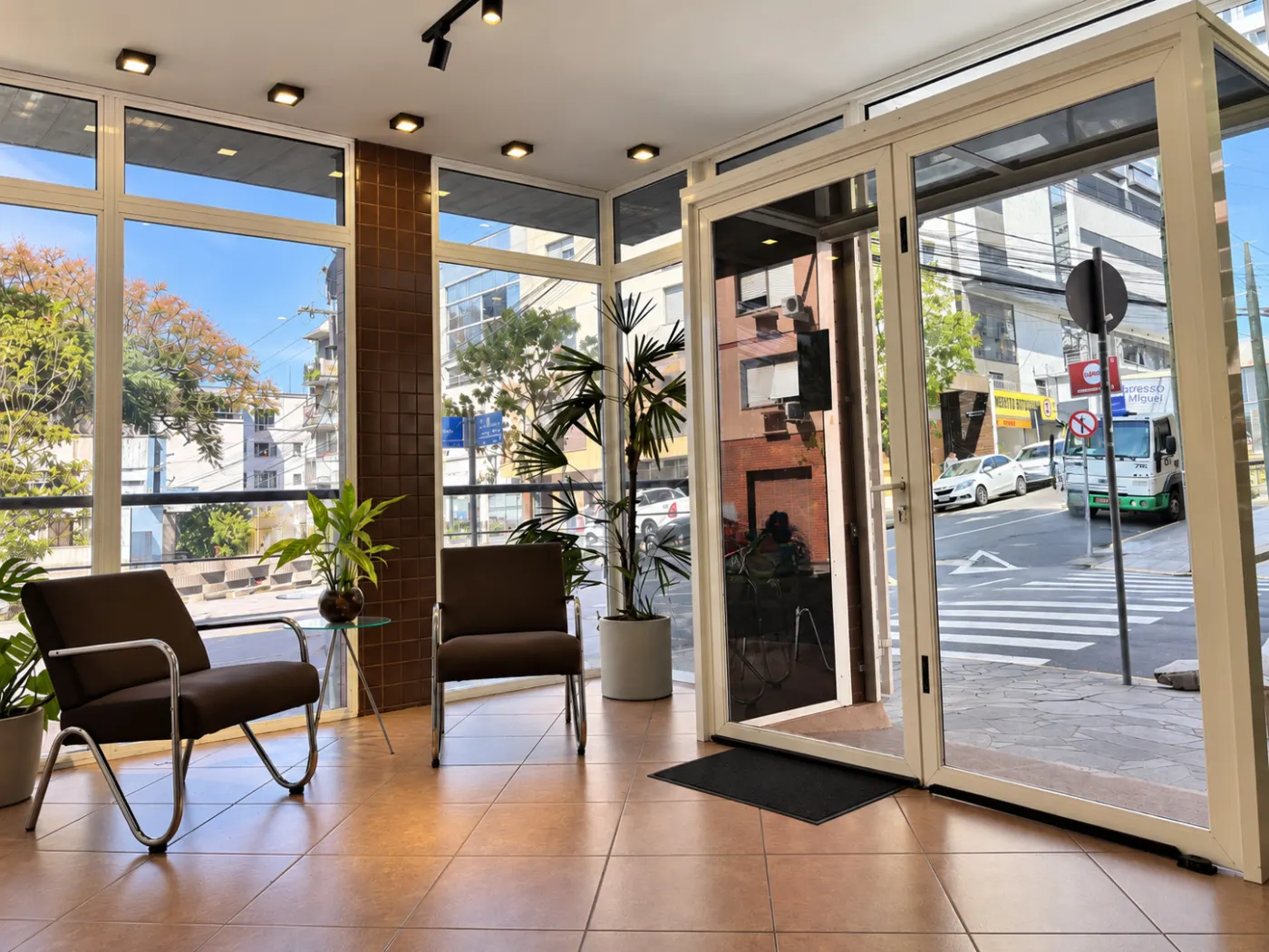 Entrada do escritório no térreo, com porta de vidro, poltronas, plantas e piso claro
