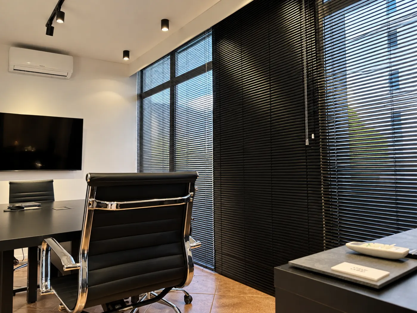 Sala de atendimento com mesa escura, cadeira executiva de couro preto, televisor na parede e persianas pretas nas janelas